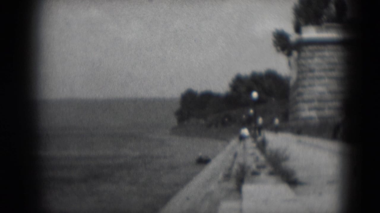 1975 8mm home-movie frame of a waterfront promenade in Middletown, Pennsylvania, with open water, lamp posts, trees and distant walkers.