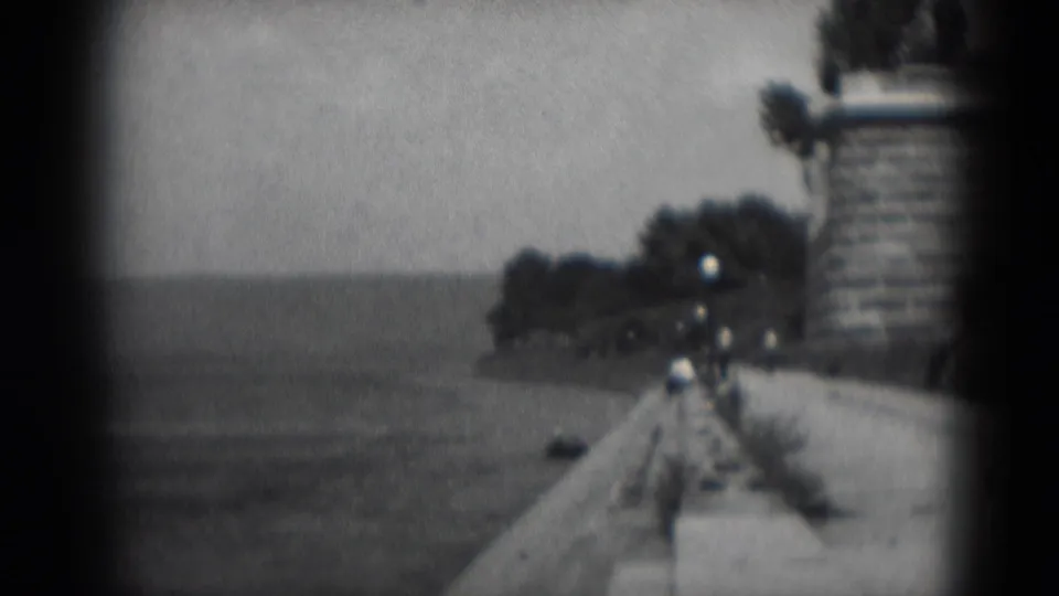 1975 8mm home-movie frame of a waterfront promenade in Middletown, Pennsylvania, with open water, lamp posts, trees and distant walkers.