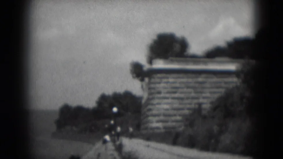 Soft-focus 1975 home-movie frame of a waterfront promenade in Middletown, Pennsylvania, with water, stonework, trees and distant pedestrians.