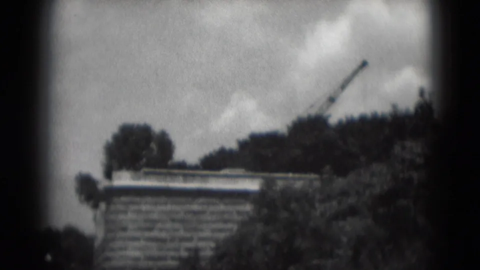 1975 home-movie frame of an older brick building, dark trees, and a distant crane under a cloudy Pennsylvania sky.