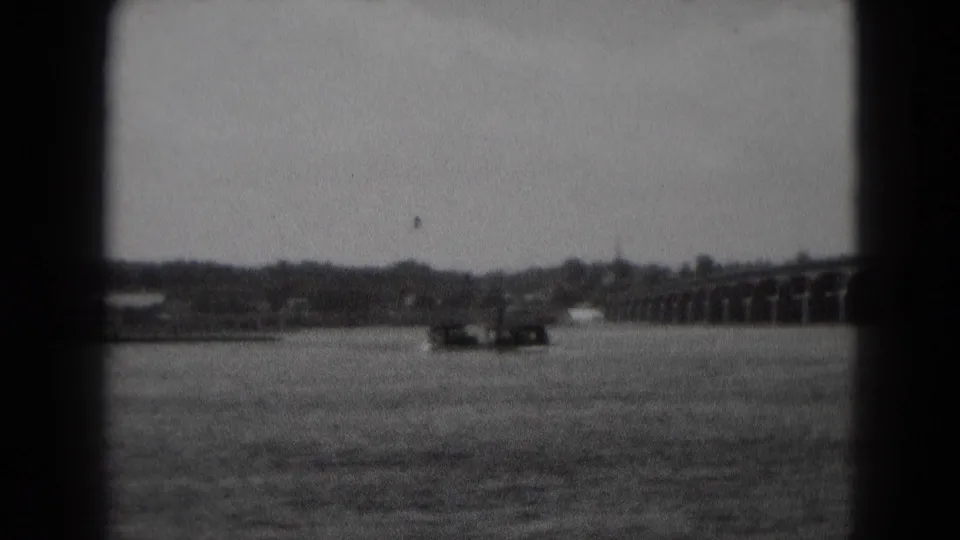 Black-and-white 1975 home-movie frame from Middletown, Pennsylvania, showing a distant boat on a river near an arched bridge.