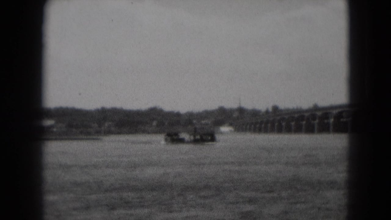 A long low boat moves across a broad river toward a multi-arch bridge in grainy black-and-white 8mm footage.