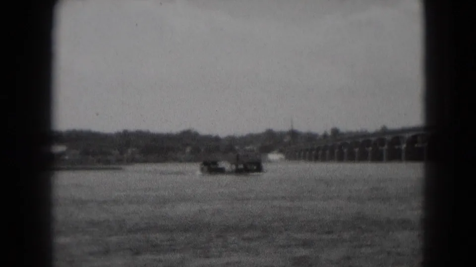 A long low boat moves across a broad river toward a multi-arch bridge in grainy black-and-white 8mm footage.