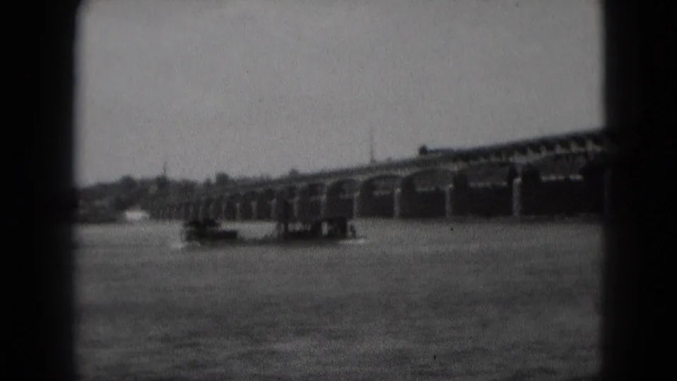 Black-and-white frame showing a boat passing on the river below the long arched bridge near Middletown, Pennsylvania.