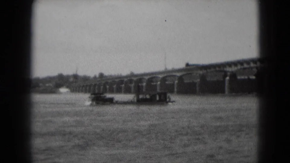 1975 8mm frame from Middletown, Pennsylvania, showing a paddle boat on rippled water near a long arched bridge.