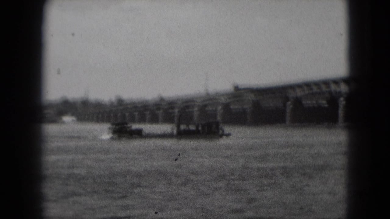 Paddle boat and wake on a broad river below an arched bridge.