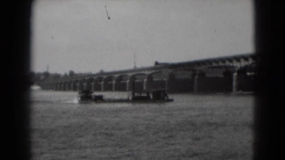 1975 home-movie frame of a paddle boat on a Pennsylvania river, with bridge arches and shoreline visible beyond.
