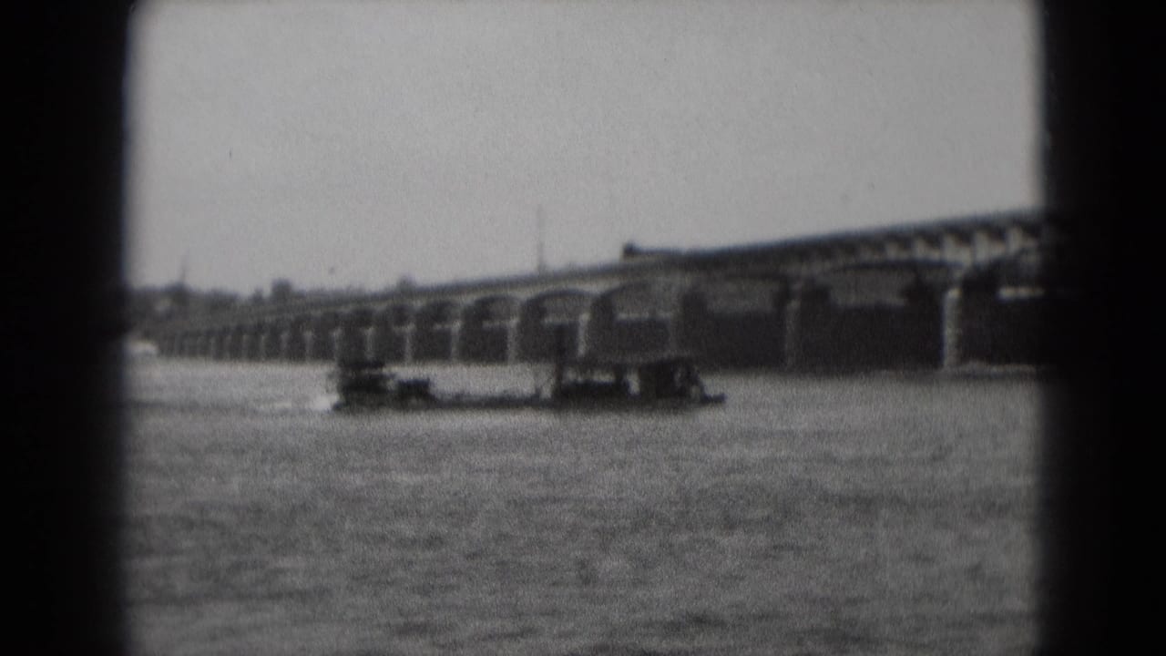 Small barge moving on a wide river below a long multi-arch stone bridge.