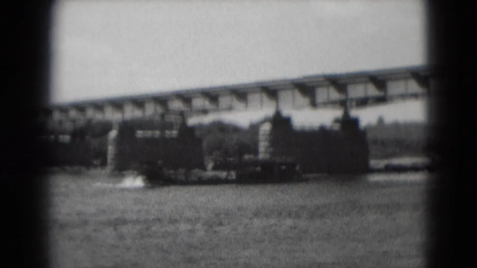 1975 8mm home-movie view of a sternwheeler-style riverboat framed by water, bridge supports, and a distant shoreline.