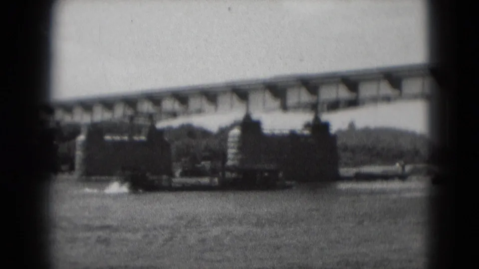 Black-and-white 8mm frame showing the sternwheeler-style boat progressing along the river under a long bridge in Pennsylvania.