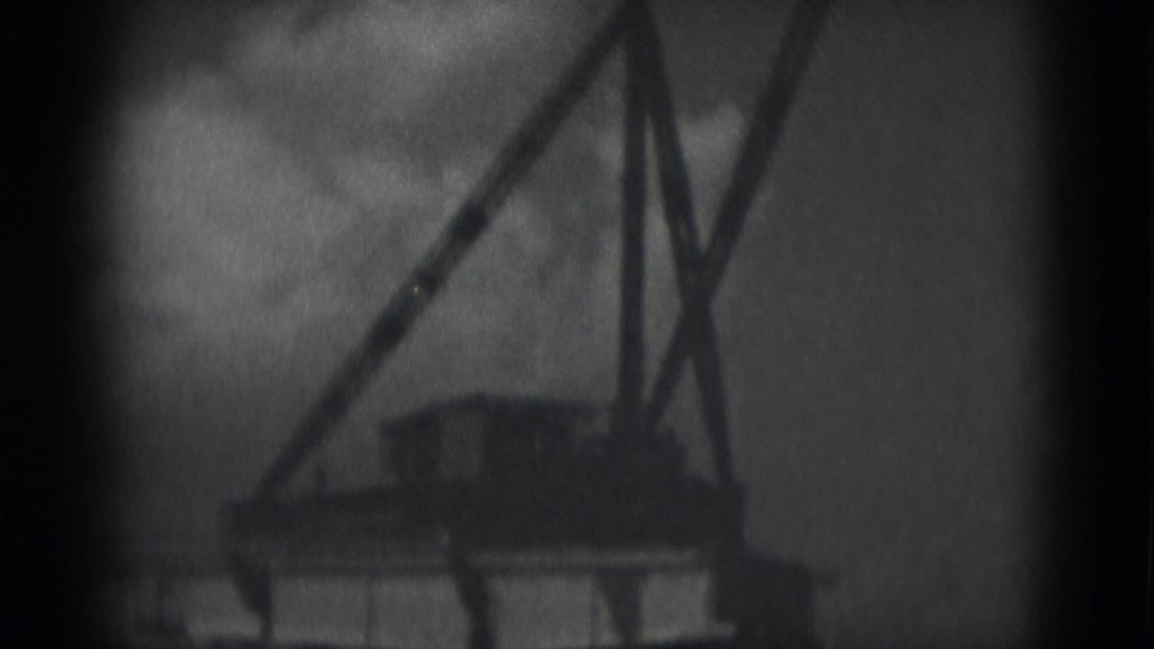 Tall A-frame or crane-like structure above low industrial forms under a cloudy sky.