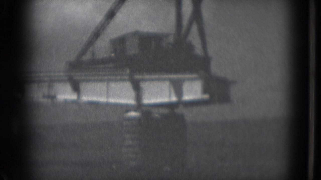Bridge segment suspended from a crane with dark ground or water below.