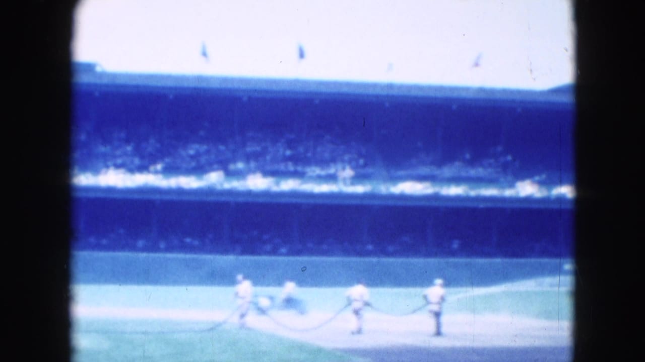 1975 Middletown baseball footage showing distant players or groundskeepers in white on the field with a crowded grandstand beyond.