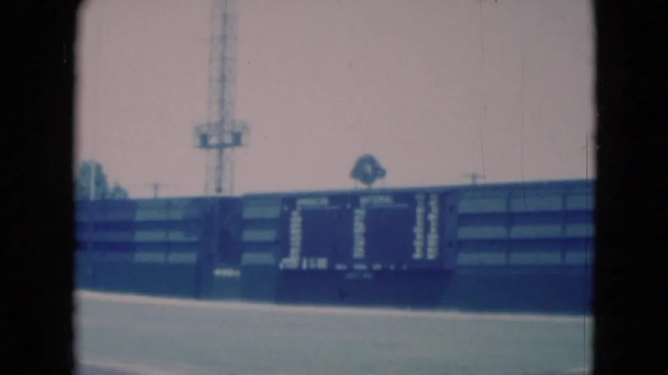1975 home-movie frame of a Middletown baseball field, centered on the scoreboard, outfield wall, and tall light tower.