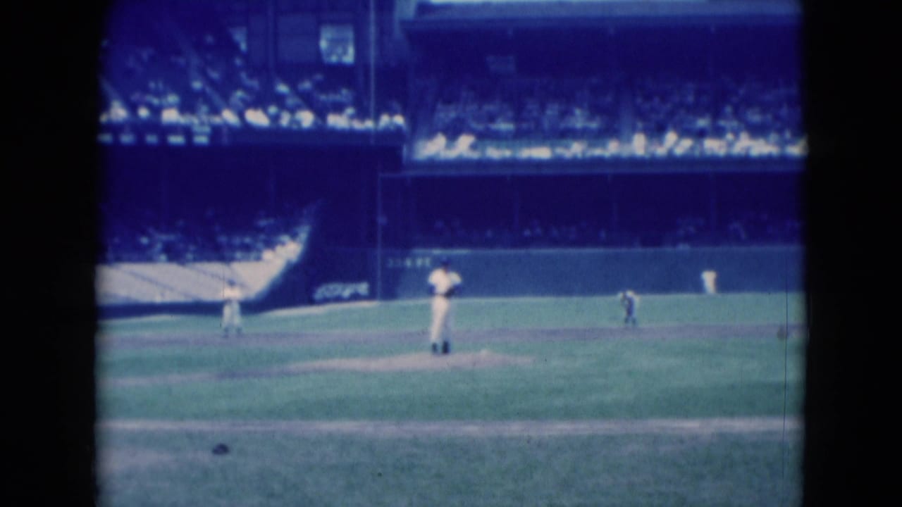 Pitcher stands on the mound with fielders and spectators visible in the stands behind him.