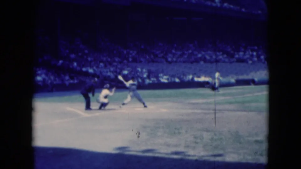 Soft 8mm frame of a 1975 Middletown baseball game with batter, catcher, umpire, and crowded stands visible.