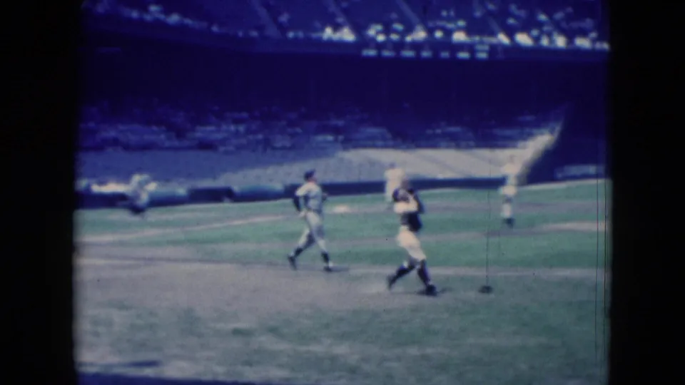 1975 Middletown baseball footage showing players crossing the grass and dirt field, framed by stadium seating and blue-tinted 8mm film borders.