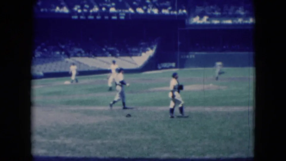 Home-movie frame of a 1975 baseball game in Middletown, Pennsylvania, with several players spread across the field and spectators in the stands.