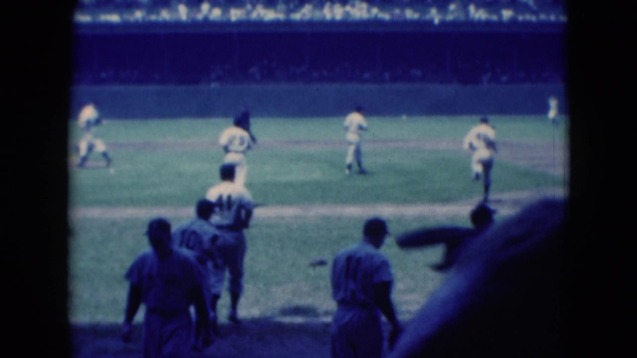 Baseball players in white uniforms move across the infield as spectators fill the stands.