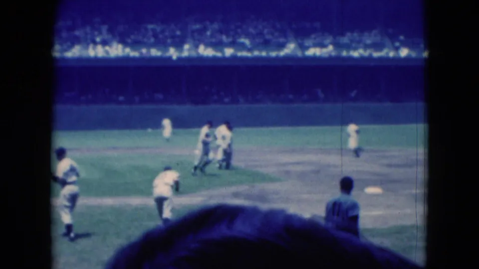 Blue-tinted 8mm frame of a 1975 Middletown baseball game with players gathered on the infield and spectators beyond.