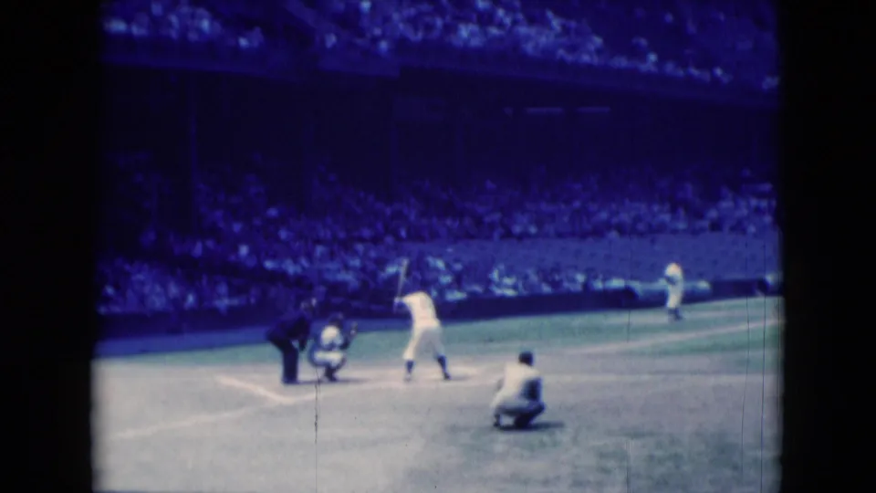 A 1975 8mm frame shows the batter set near home plate as the catcher crouches, the umpire leans in, and the crowd fills the stands.