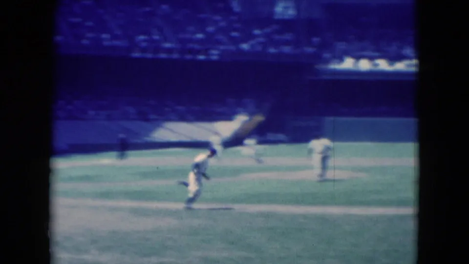 Blue-tinted 8mm frame from the 1975 Middletown baseball game showing a player bending low on the grass and infielders positioned nearby.