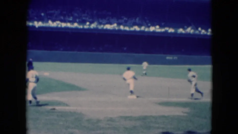 1975 Middletown, Pennsylvania 8mm frame showing baseball players in white uniforms on an infield before a crowded stadium backdrop.