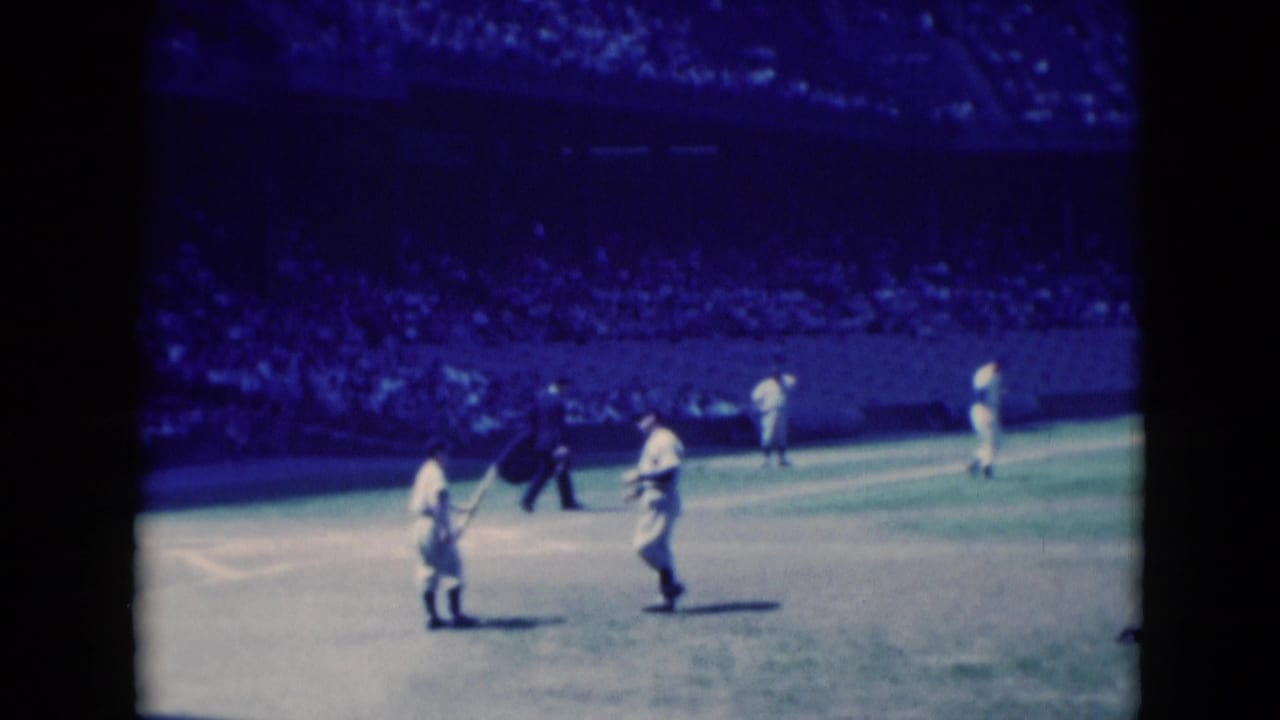 Players in white uniforms stand on a bright playing field with a crowded stand behind them.