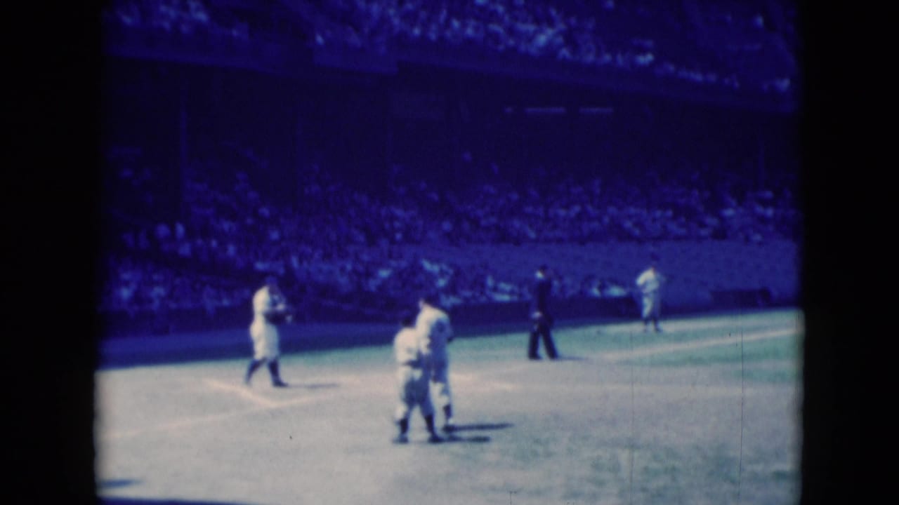 1975 Middletown, Pennsylvania home-movie frame of cricket players gathered on the field with a dark-clothed official and spectators beyond.