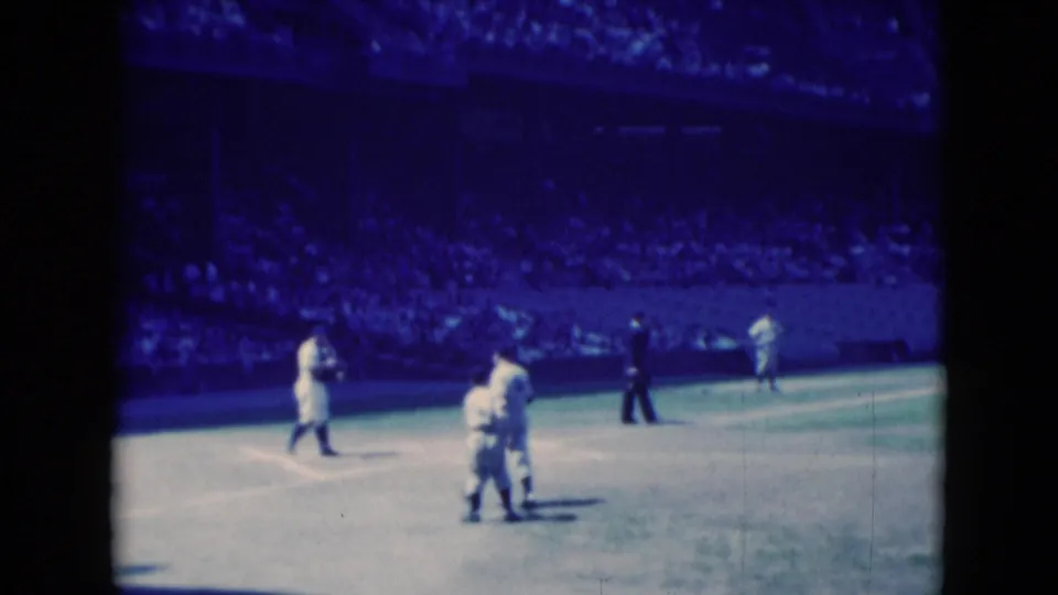 1975 Middletown, Pennsylvania home-movie frame of cricket players gathered on the field with a dark-clothed official and spectators beyond.