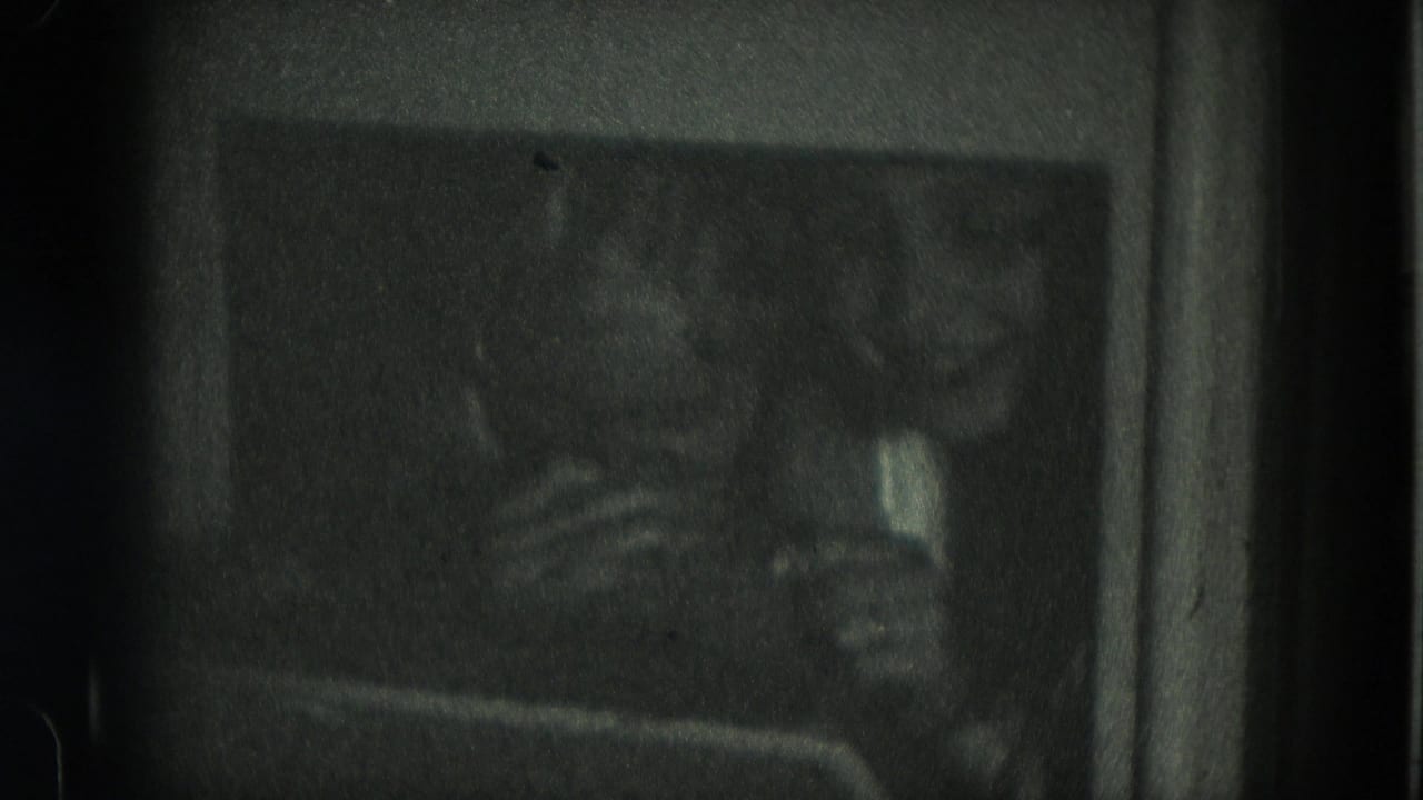 Two girls appear in a dim 1975 Milwaukee 8mm home-movie interior, their faces soft and partly obscured by low light.