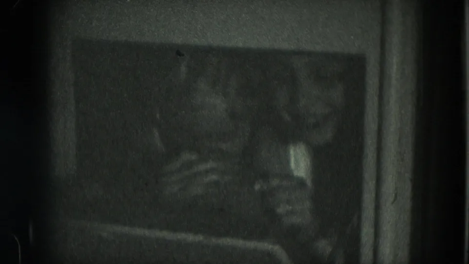 Two girls appear in a dim 1975 Milwaukee 8mm home-movie interior, their faces soft and partly obscured by low light.