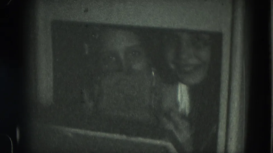 In a dark 8mm interior, two girls look toward the camera with one face clearer and the other receding into shadow.