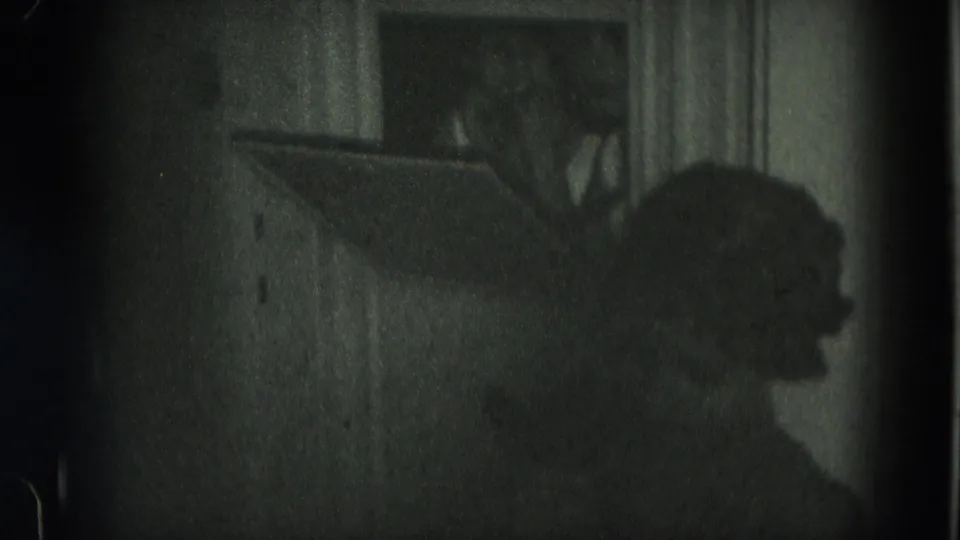 A dark interior frame shows a person in profile near a wall or window, with details obscured by shadow and grain.