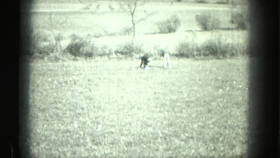 A bright 8mm outdoor field scene shows two small figures bending in grass, with bare trees and brush in the background.