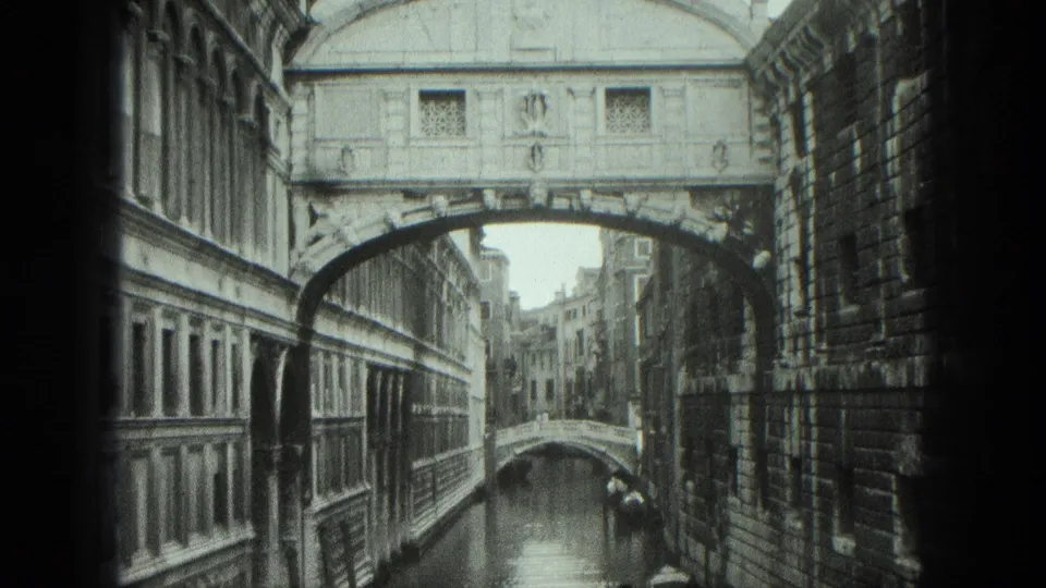 1975 Venice frame showing the Bridge of Sighs above canal water, bordered by tall stone buildings and a distant canal bridge.
