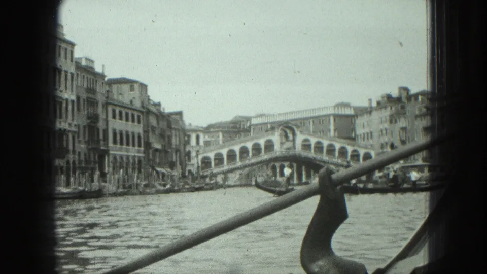 1975 8mm Venice view toward the Rialto Bridge from a moving boat, with canal water and gondola hardware crossing the frame.