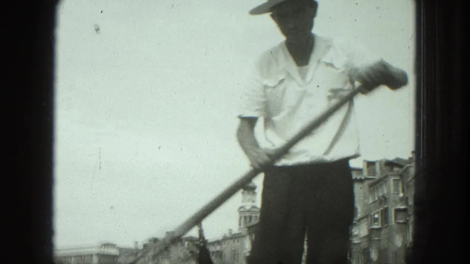 1975 Venice 8mm frame of a gondolier in a light shirt and straw hat working an oar, with canal architecture below the horizon.