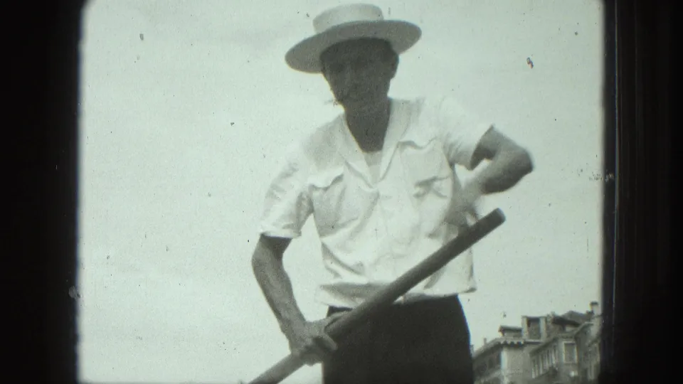 1975 Venice home-movie frame showing a gondolier in a straw hat holding an oar against a pale sky and canal-side buildings.