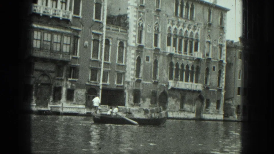 1975 Venice frame with a gondolier propelling a gondola past tall canal-side buildings and arched windows.