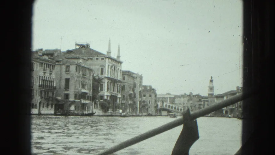 1975 Venice frame looking along the canal toward buildings, a bridge, and distant vertical landmarks from a moving viewpoint.