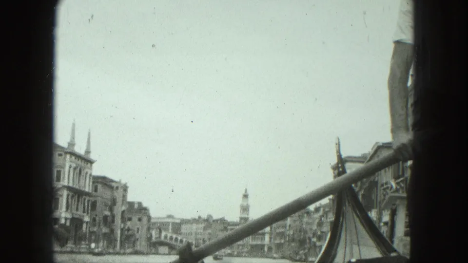 1975 Venice gondola perspective with the prow at right, a rowing pole across the frame, canal buildings, and a distant bell tower.