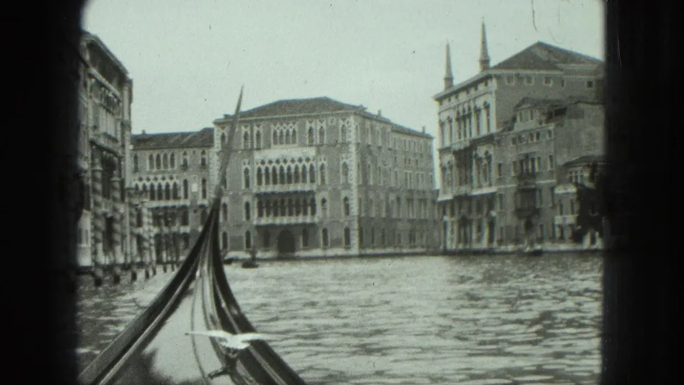 1975 Venice canal frame with gondola prow in the foreground, rippled water, and ornate waterfront buildings beyond.