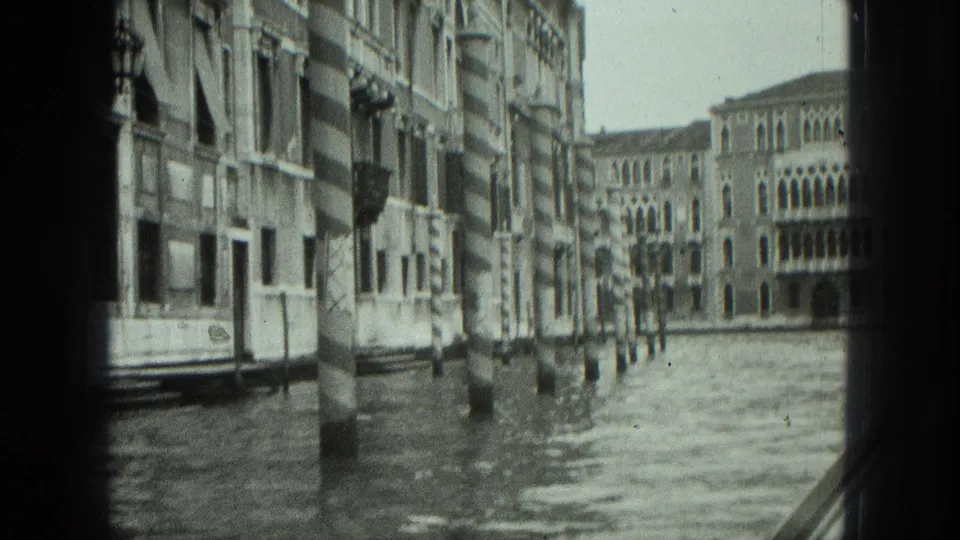 1975 Venice canal view from a moving boat, framed by dark edges with historic buildings and striped mooring poles along the water.