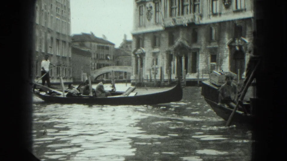 1975 Venice canal view with a gondolier standing in a gondola near ornate building facades and a small bridge.