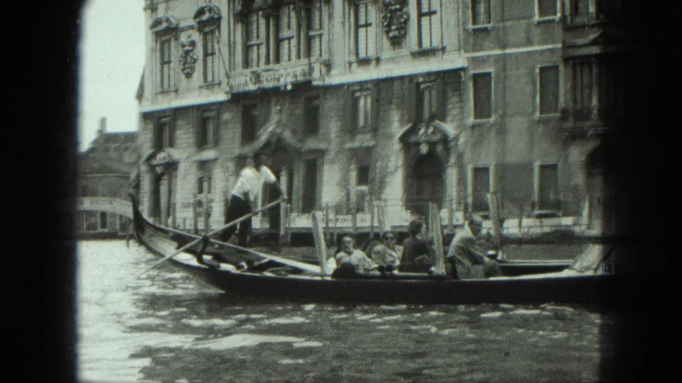 1975 Venice home-movie frame showing a gondolier standing at the stern while tourists sit in a gondola near canal-front architecture.