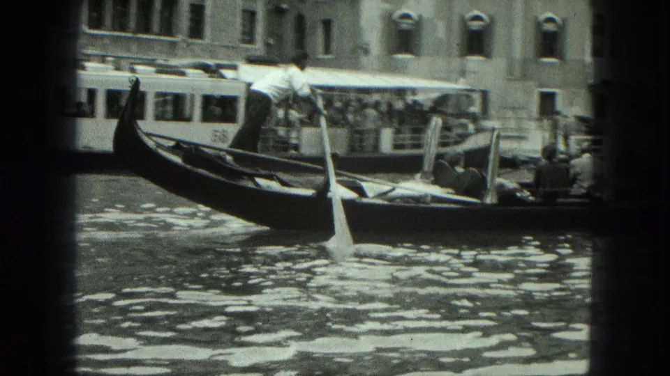 1975 Venice 8mm frame showing a gondola crossing the canal with a motorboat behind it and tourists seated near the center of the boat.