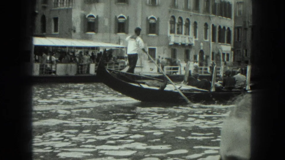 1975 Venice frame showing the gondolier standing in the gondola as passengers ride past moored boats and canal-front buildings.
