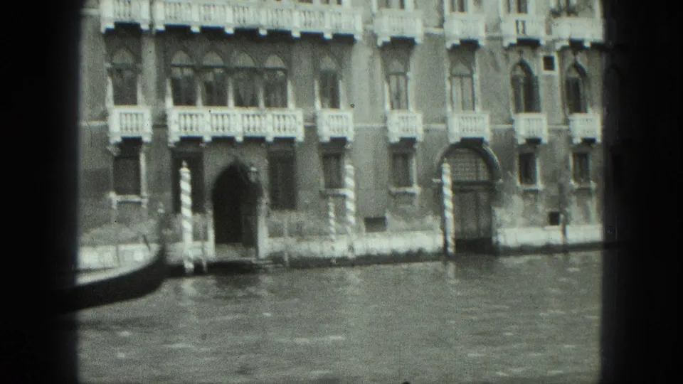 Black-and-white 8mm view of a Venetian waterfront facade with balconies, arched windows, and canal water.
