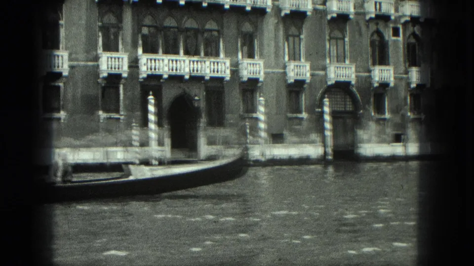 1975 Venice home-movie frame with a gondola bow, canal water, balconies, and arched waterfront entrances.